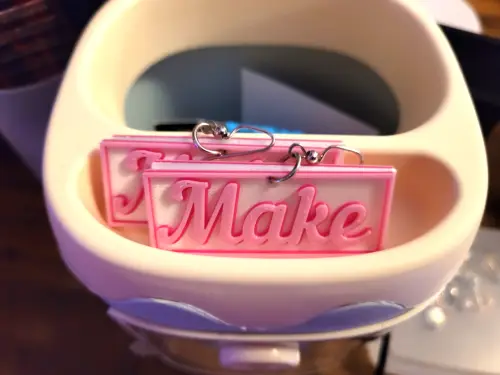 a close up of a rectangular earring that says 'Maker' on it in the style of the Barbie movie font and colors.