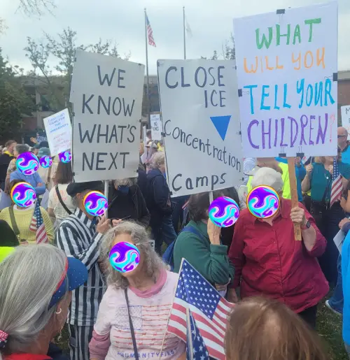 A protester is carrying a sign that says 'CLOSE ICE CONCENTRATION CAMPS' and a protester behind her is wearing a striped concentration camp outfit, holding a sign that says, 'WE KNOW WHAT'S NEXT.' Another protester beside them has a sign: 'WHAT WILL YOU TELL YOUR CHILDREN?'